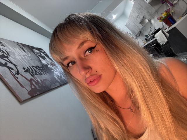 SweetMirakle has long blonde hair, brown eyes, and a nose piercing. She's wearing a white tank top with pink lips and a small cross necklace.