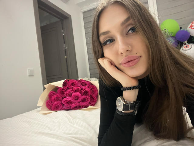 SweetMirakle with long brown hair, nose ring, and lip piercing, resting her head on her hand, wearing black, showing pink roses on a bed, and a wristwatch.