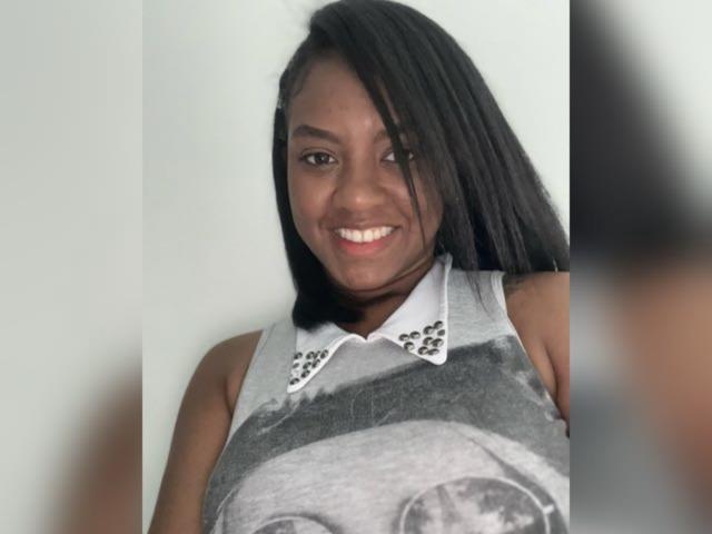 TiffanySammy has dark skin, straight black hair, and a big smile showing white teeth. She's wearing a gray sleeveless top with a white collar and small studs.