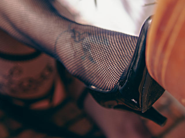 Elizabeth Duponts schwarzer Fuß, bedeckt mit Fischgrätmuster-Strümpfen, in einem glänzenden schwarzen Stiletto-Absatz, wobei ein Tattoo an ihrem Bein zu sehen ist. Der Fokus liegt auf ihrem Absatz und der Textur der Fischgrätmuster-Strümpfe..