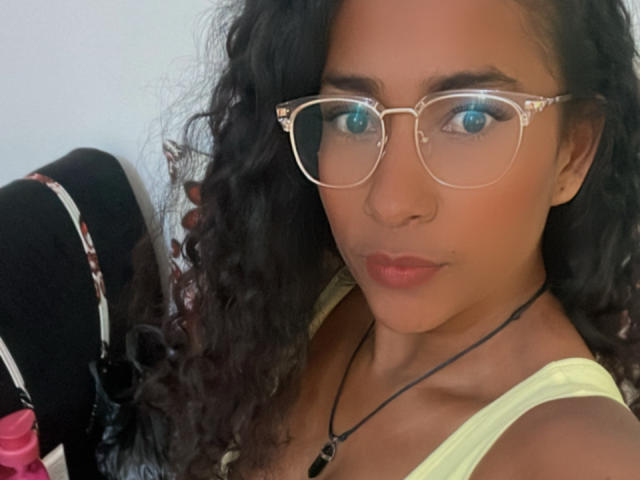 Thannia has curly black hair, blue eyes, and wears clear glasses with a yellow tank top, showing a cute face with pink lips and a black necklace.