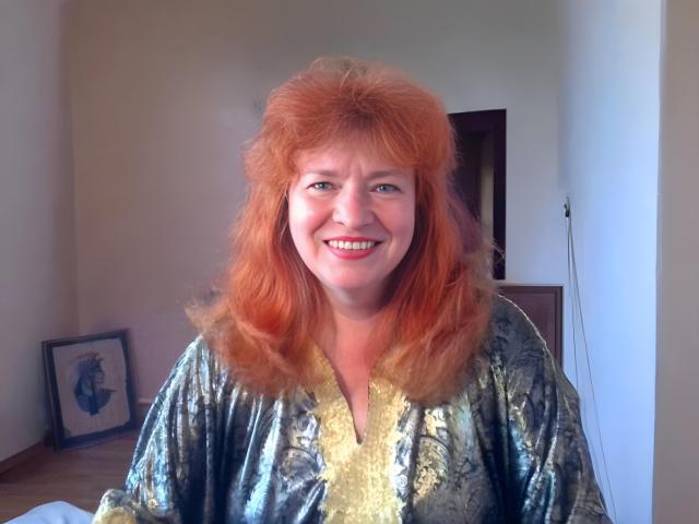 LadaMay with red, curly hair, smiling, wearing a shiny, patterned, gold-trimmed robe, showing her tits slightly, in a photo.