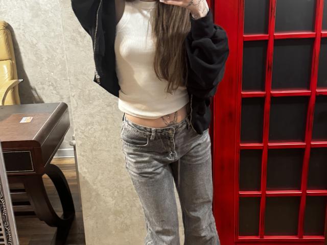 SensualMila with long brown hair, wearing a black jacket, white tee, and gray jeans, showing a small tattoo on her lower right belly, holding a phone in front.