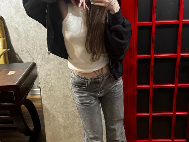 SensualMila has long brown hair, a white tank top, black hoodie, and grey jeans. She's showing a tattoo just above her jeans' waistband. Her phone covers her face.