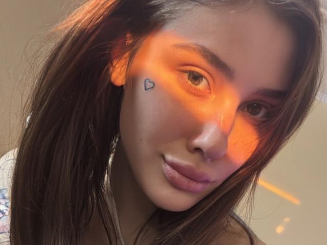 SensualMila with long brown hair, orange light on her face, small heart tattoo under her right eye, and slightly pouty pink lips, looking into the camera.