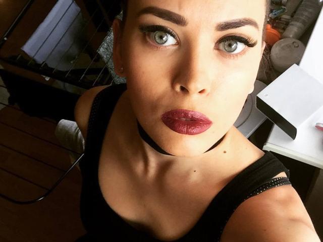 AlicexFresh with green eyes, dark red lipstick, black choker, black top, and raised eyebrows, looking up at the camera with a slight pout. Her tits are visible.