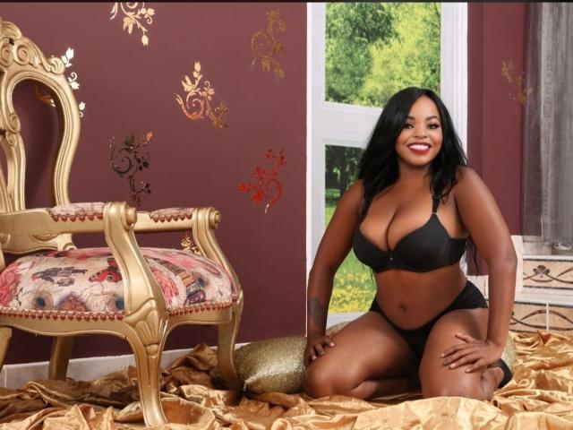 TatianaMarin, dark-skinned with black hair, kneeling in black bra and panties, showing her big tits and ass, smiling, on gold satin with ornate chair and window.
