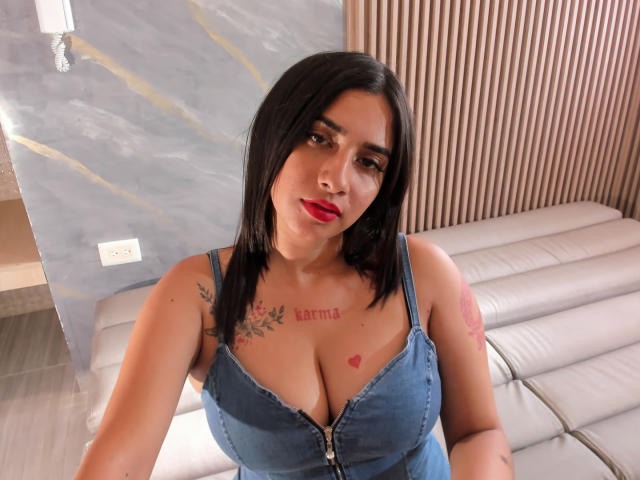 SofiTattoo with black hair, red lipstick, and blue denim dress, showing her big tits and tattoos, including "Bartia" and a heart on her chest, sitting on a white couch.