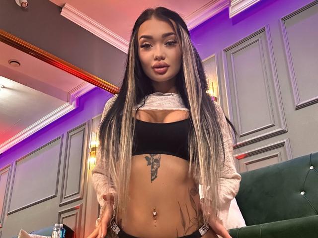SashaFerrera has long, straight blonde and black hair, wearing a black bra, showing her tits, belly button piercing, tattoo under her tits, and a grey sweater.