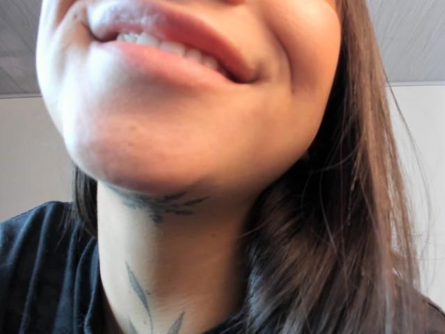 KayleeM's face, showing her slightly parted pink lips and a small tattoo on her neck with a subtle smile. Brown hair, black shirt, light skin. Close-up shot.