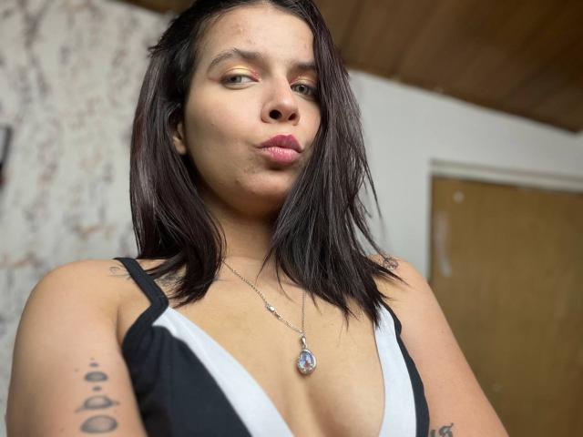 KaylinJulithe with medium tits in a black and white dress, pouty lips, dark shoulder-length hair, tattoo on right arm, necklace with blue gem, wooden ceiling background.