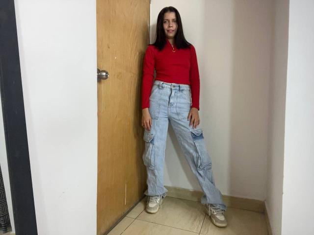 KaylinJulithe, con cabello castaño oscuro, una camisa roja de manga larga, pantalones cargo azul claro y zapatillas blancas, está de pie frente a una puerta de madera, mirando directamente hacia adelante, con una expresión neutra..
