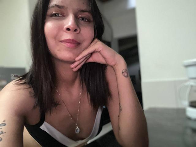 KaylinJulithe with black hair, light brown skin, small tits, wearing a black and white top, hand on chin, subtle tattoos, necklace, and a slight smile in a selfie.