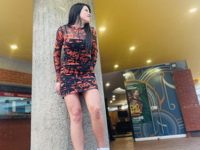 AmelieMuller standing against a gray concrete pillar, wearing a tight, black and orange patterned dress, showing her ass and pussy, with long black hair, looking to the side.