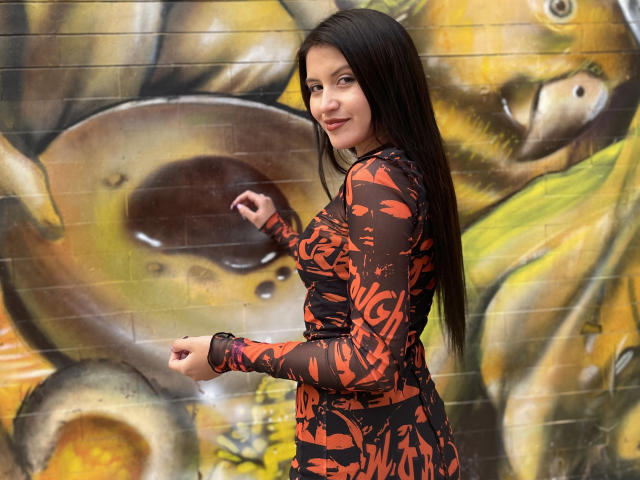 AmelieMuller with long black hair, smiling, wearing a tight orange and black patterned dress, standing in front of graffiti, pointing with her right hand, showing her tits.