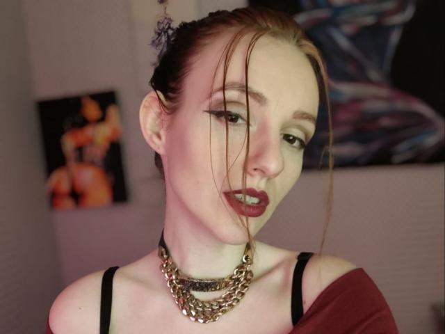 KatKitty with fair skin, dark red lipstick, and a chain necklace, has brown hair in a messy updo, black bra, and a seductive expression, with a blurred background.