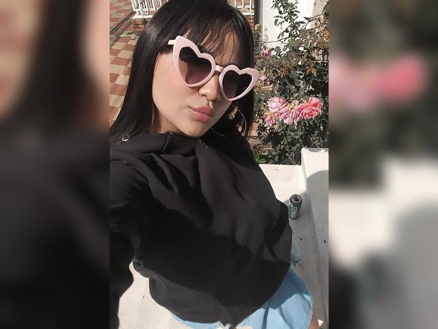 Karina Blake trägt herzförmige, pinke Sonnenbrillen, eine schwarze Kapuzenjacke und blaue, hochgeschnittene Hosen. Sie zieht die Lippen zusammen, und ihr dunkles Haar und ihre helle Haut bilden einen Kontrast zu ihrem Outfit..