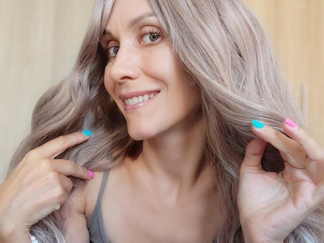 EmieFlower with long, wavy, grayish-blonde hair, smiling, showing pierced bottom lip, gray tank top, pink and blue nails, pointing at hair, light skin, cute.