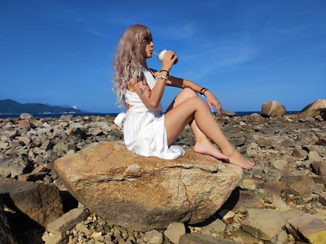 EmieFlower with long, wavy blonde hair, sitting on a rock, wearing a white dress, holding a shell, barefoot, on a rocky beach, clear blue sky.