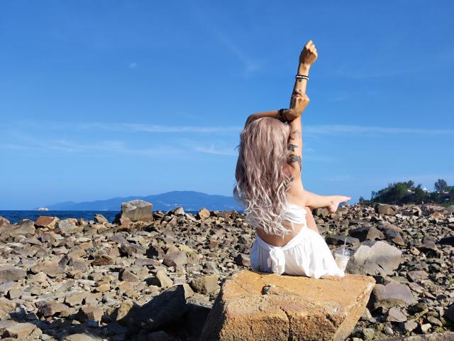 EmieFlower with pink wavy hair, arms raised, wearing a white dress, sitting on rocks, blue sky, ocean in background, tattoos on arm, sunny day, rocky shoreline.