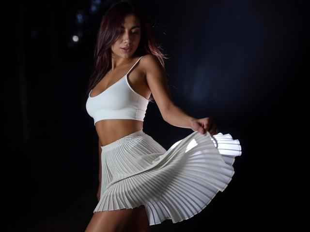 MindyDesire with white crop top and pleated skirt, flapping skirt, toned arms, brown hair, dark background, small tits, white bra, sexy, attractive.