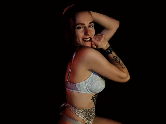 KatKitty with pale skin, blue lace bra, and silver lace panties, showing tattoos, smiling, biting finger, dark background, right arm raised. Boobs, tits, and pussy not visible.
