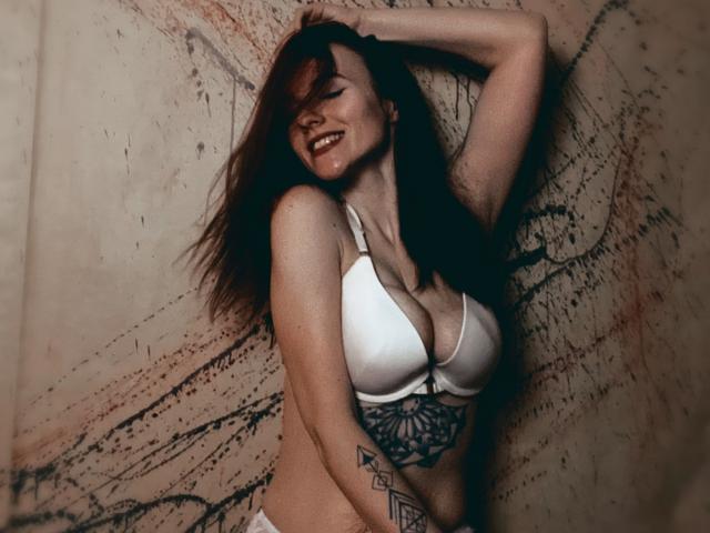 KatKitty, con lunghi capelli castani, sorridente, indossa un reggiseno bianco, mostrando il seno, e rivela un tatuaggio sotto il seno. È appoggiata a un muro con una superficie irregolare, con una mano tra i capelli..