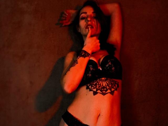 KatKitty with black lace bra, tattoo on chest, hand over mouth, red lighting, shadow on wall, black lace panties, arms above head, sexy, confident, dark hair, slightly open mouth.