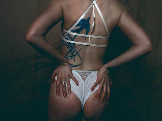 KatKitty's bare back shows a blue bird tattoo, wearing white strappy lingerie with her hands on her butt, fingers with a ring. Her ass is visible, looking sexy.