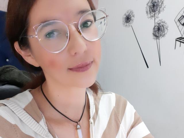 AntonnellaS with glasses, light skin, brown hair, pinkish lipstick, wearing a striped shirt, black necklace, subtle smile, green eyes, dandelion wall art, and a blue chair.