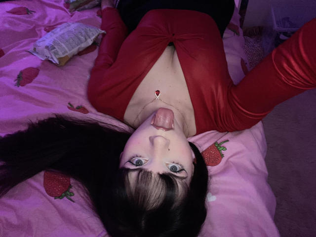 JennyMay, con cabello negro, una blusa roja y un collar, con la lengua afuera, acostada en una cama rosa con forma de fresa, ojos azules, piel blanca y un escote visible. Sus brazos están levantados..