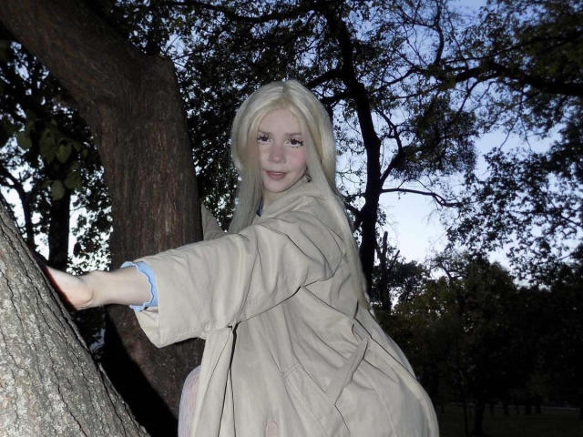 JennyMay, avec des cheveux blonds, une peau claire, portant un manteau beige, appuyée contre un arbre, souriante, montrant un petit postérieur, manches à rayures bleues, nuit, branches d'arbres en arrière-plan..