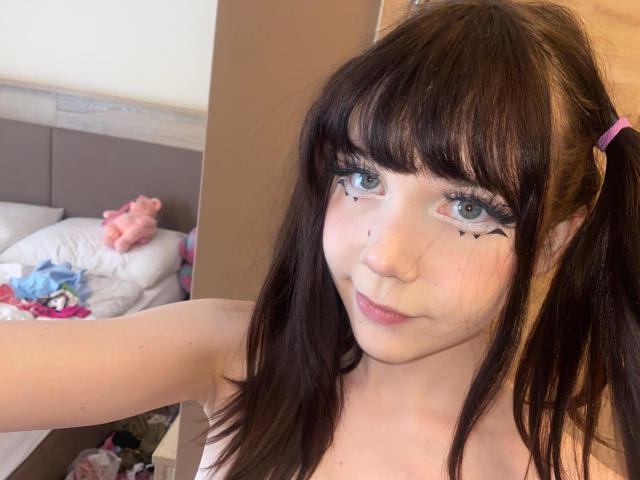 JennyMay, avec des couettes, des yeux bleus, du crayon noir pour les yeux, un anneau au nez, une peau claire, de petites seins, et un léger sourire, prenant un selfie dans une chambre..