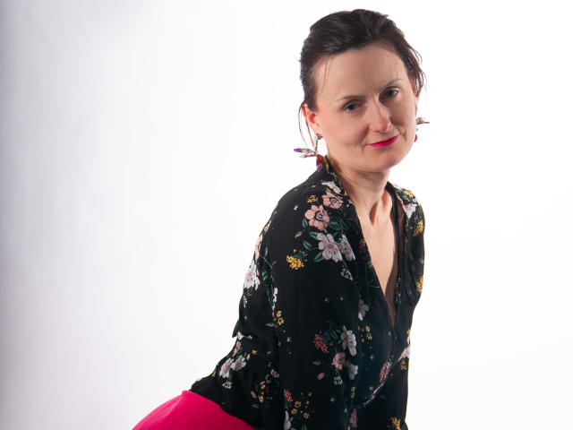PattyAmore mit einer dunkelblauen Bluse mit Blumenmuster, einem pinken Rock und rotem Lippenstift, wirkt leicht genervt, trägt bunte Ohrringe, vor einem weißen Hintergrund..