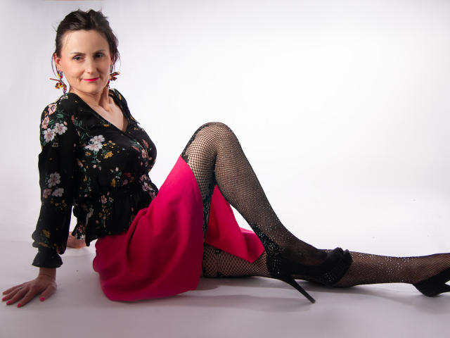 PattyAmore mit einer blumigen schwarzen Bluse, einem pinken Rock, Fischernetzstrümpfen und schwarzen High Heels, sitzend auf einem weißen Boden, Beine ausgestreckt, lächelnd, rote Ohrringe, mittelgroße Brüste, süß..