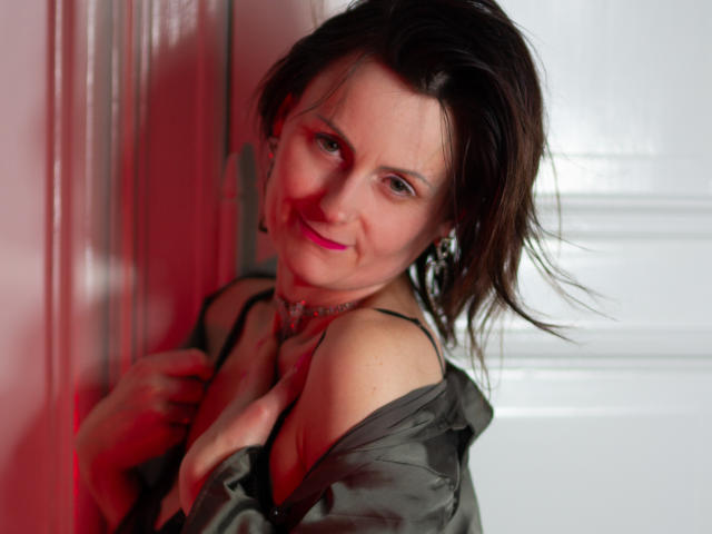 PattyAmore with messy brown hair, pink lipstick, and a sly smile, wearing a green shirt, black bra strap, and choker, leaning against a red-lit wall.