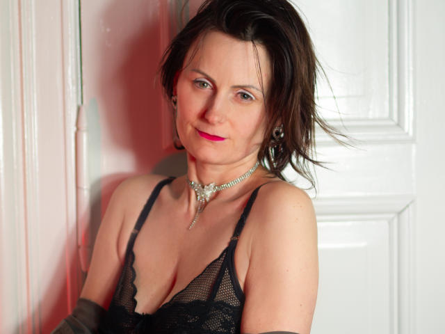 PattyAmore with dark hair, pink lipstick, black lace bra, and necklace, leaning against white door, smiling slightly, showing small tits and a cute face.