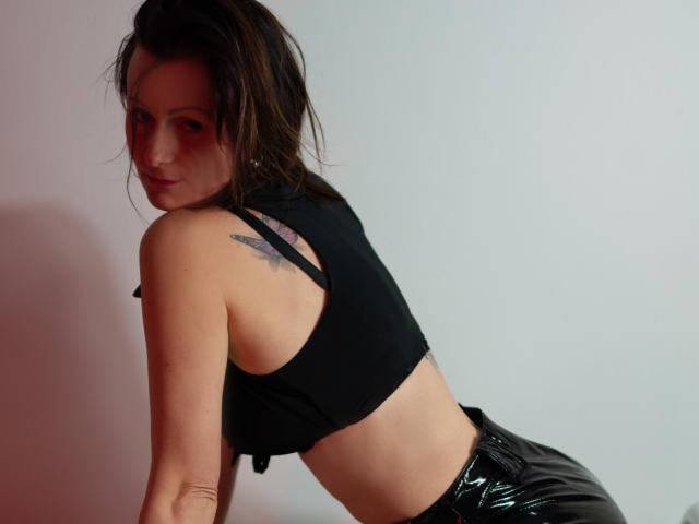PattyAmore com maquiagem vermelha nos olhos, cropped top preto e calças pretas brilhantes, inclinada para frente, mostrando uma tatuagem no ombro direito e olhando por cima do ombro..