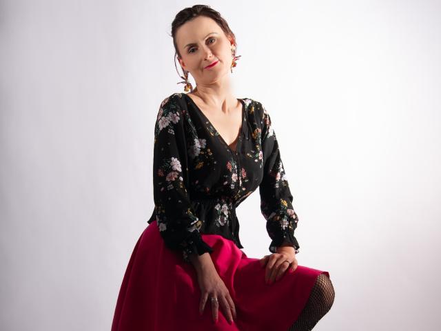 PattyAmore con una blusa floral oscura, falda roja y media de rejilla, sentada, sonriendo, con las manos en la falda, pechos medianos, fondo blanco, aretes rojos, cabello castaño, piel clara..