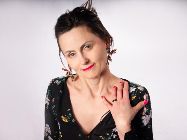 PattyAmore mit dunklem Haar, einem schwarzen Kleid mit Blumenmuster, rosafarbenen Nägeln und Schmetterlings-Ohrringen, die leicht lächelt, mit der Hand in der Nähe des Halses, rosafarbenem Lippenstift, einem weißen Hintergrund und dezentem Make-up..