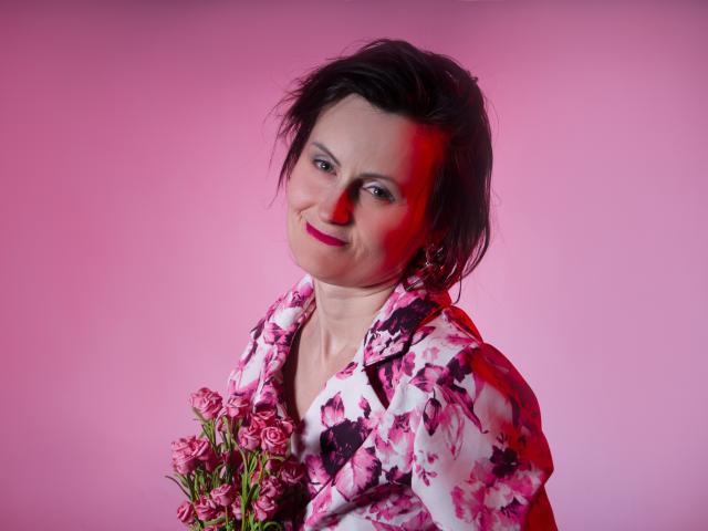PattyAmore, con rosas rosadas, lleva una camisa floral rosa y blanca, tiene el pelo corto y negro, lápiz labial rojo y una sonrisa encantadora, sosteniendo flores frente a ella..