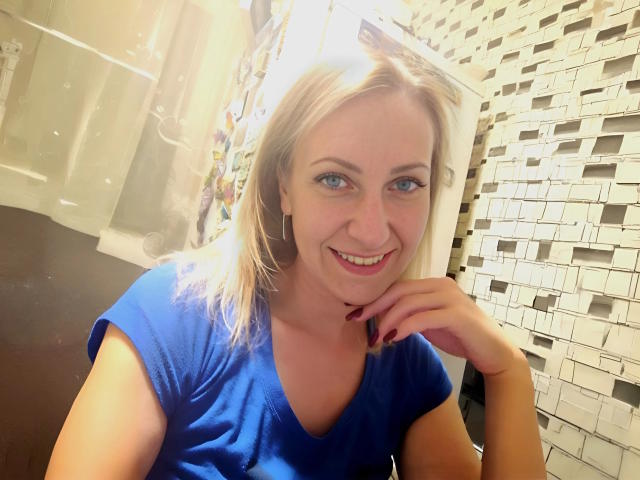 Blonde AshleyXHoney mit blauem Hemd, lächelnd, helle Haut, blaue Augen, kleine Nase, roter Lippenstift, Ohrringe und rotem Nagellack, mit dem Kinn in der Hand im Badezimmer..