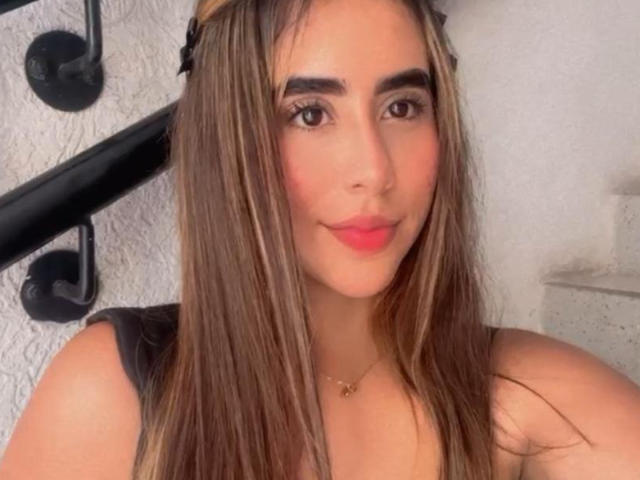 OlivaStellas, con cabello castaño largo, cejas oscuras, lápiz labial rosa y una sonrisa encantadora, viste una blusa negra, lleva un collar de oro pequeño y un accesorio para el cabello negro..
