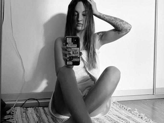 Black-and-white photo of MaryLiana with long hair, tattooed arm, sitting on floor, wearing towel, legs spread, holding phone, looking down, shadow on wall, wooden floor, rug.
