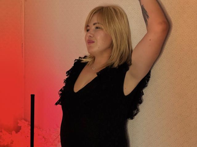 CurlyDreams with blonde hair, wearing a black ruffled top, arm raised, showing a tattoo, standing against a textured wall, red and orange lighting.