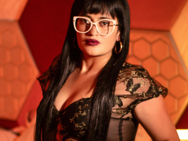 BriaVolkova with long black hair, wearing white glasses, gold hoop earrings, and a sheer black lace top, showing her tits, with dark red lipstick and a sexy gaze.