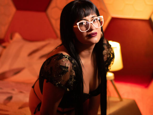 BriaVolkova with black hair and white glasses, wearing a black lace top with green embroidery, leaning forward, showing her tits, in a warmly lit bedroom.
