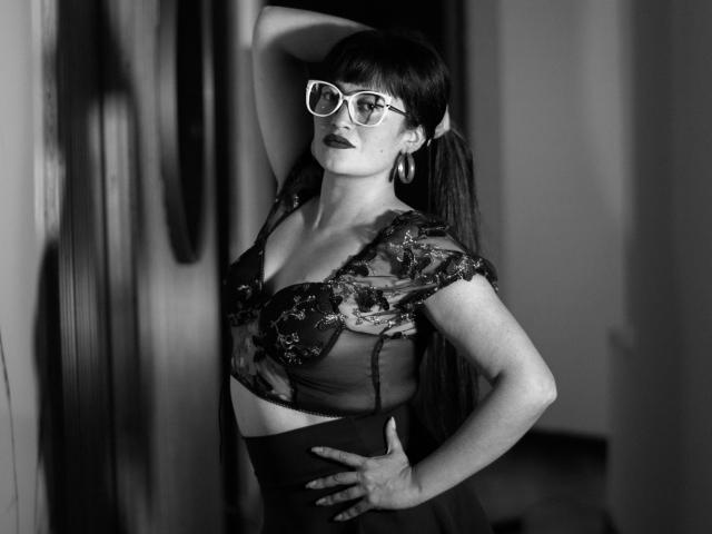 BriaVolkova with long dark hair, wearing glasses, sheer lace top, and high-waisted skirt, hand on hip, arm above head, showing her tits and dark lipstick. Black-and-white photo.