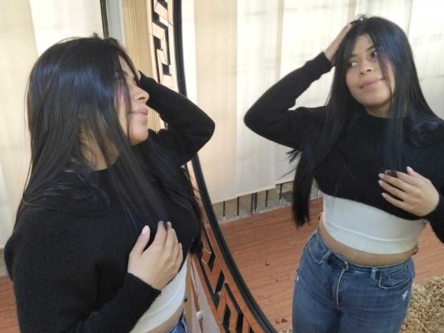 AnaTaylor, avec de longs cheveux noirs, un pull noir, un crop top blanc et un jean bleu, debout, une main dans les cheveux, une main sur la poitrine, reflet dans un miroir, souriante, montrant ses petites seins..