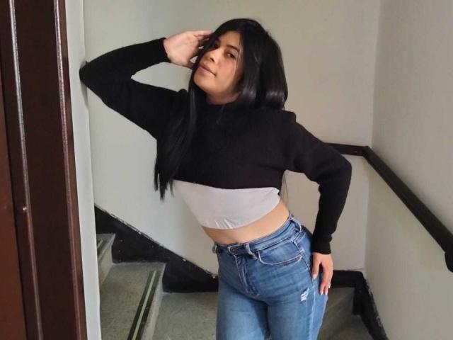 AnaTaylor con lunghi capelli neri, top corto bianco e nero, jeans blu a vita alta, in piedi sulle scale, con una mano sulla testa, che guarda di lato, ringhiera nera a destra..